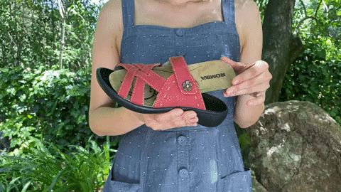 A woman wearing a blue dress installs a removable insole on a red sandal while standing in a garden.